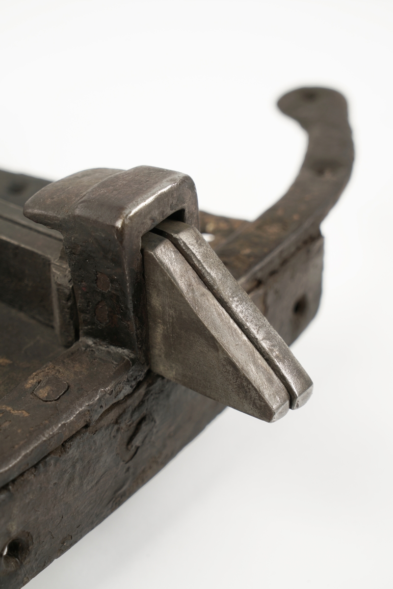 Dørlås av jern. Formet som en person, som står på en meie. Grov låsemekanisme med en hendel som gjør at låsebolten kan skyves til side, og dermed åpne døren.