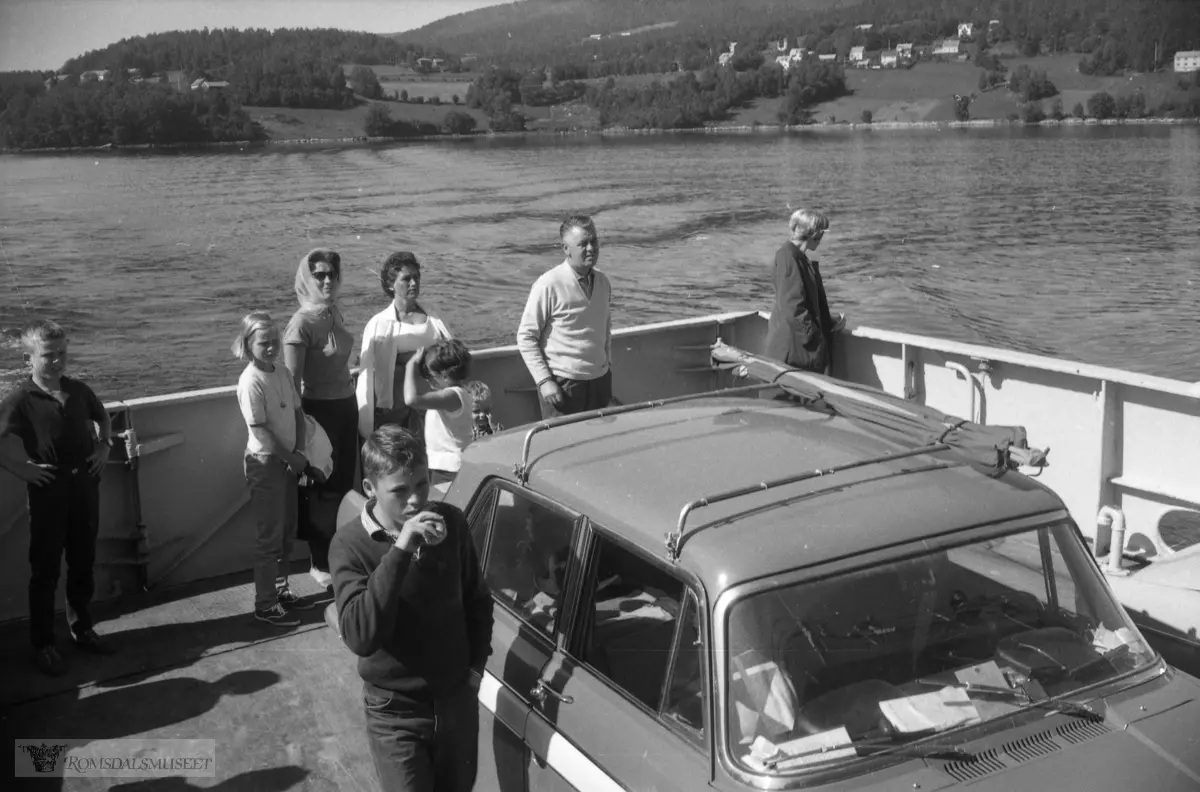 "juli 1965 Surnadal".Bildet viser akterdekket av BF Tingvoll over Tingvollfjorden på vei fra Angvik til Tingvoll. "Sørlandsveien" eller riksvei nr. 666 som går igjennom Angvik er ikke påbegynt her..Bilen er en Austin Cambridge (eller parallellen Morris Oxford). Denne versjonen, med et belte i lysere farve, var ganske ny da bildet ble tatt.