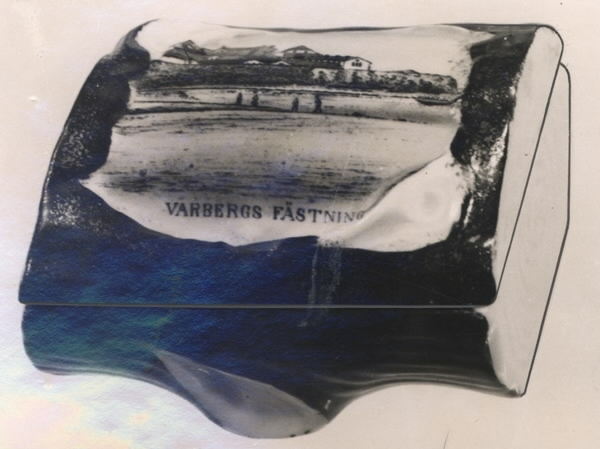 Ask med lock, porslin till förvaring av smycken. Rombisk i form av en avsågad ekstock. På locket infälld bild av "VARBERGS FÄSTNING" i färg.