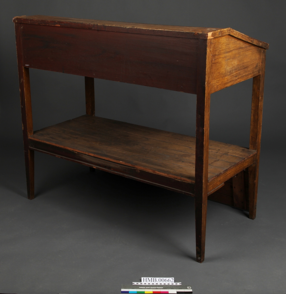 Rektangulært med skrånende skriveplate. To skuffer i front. Høye bein. Omtrent midt på beina er en hylleplate hvor det er hengslet en skapdør med en ring til håndtak. Skapdøren er hengslet i overkant og trekkes opp for å åpne.