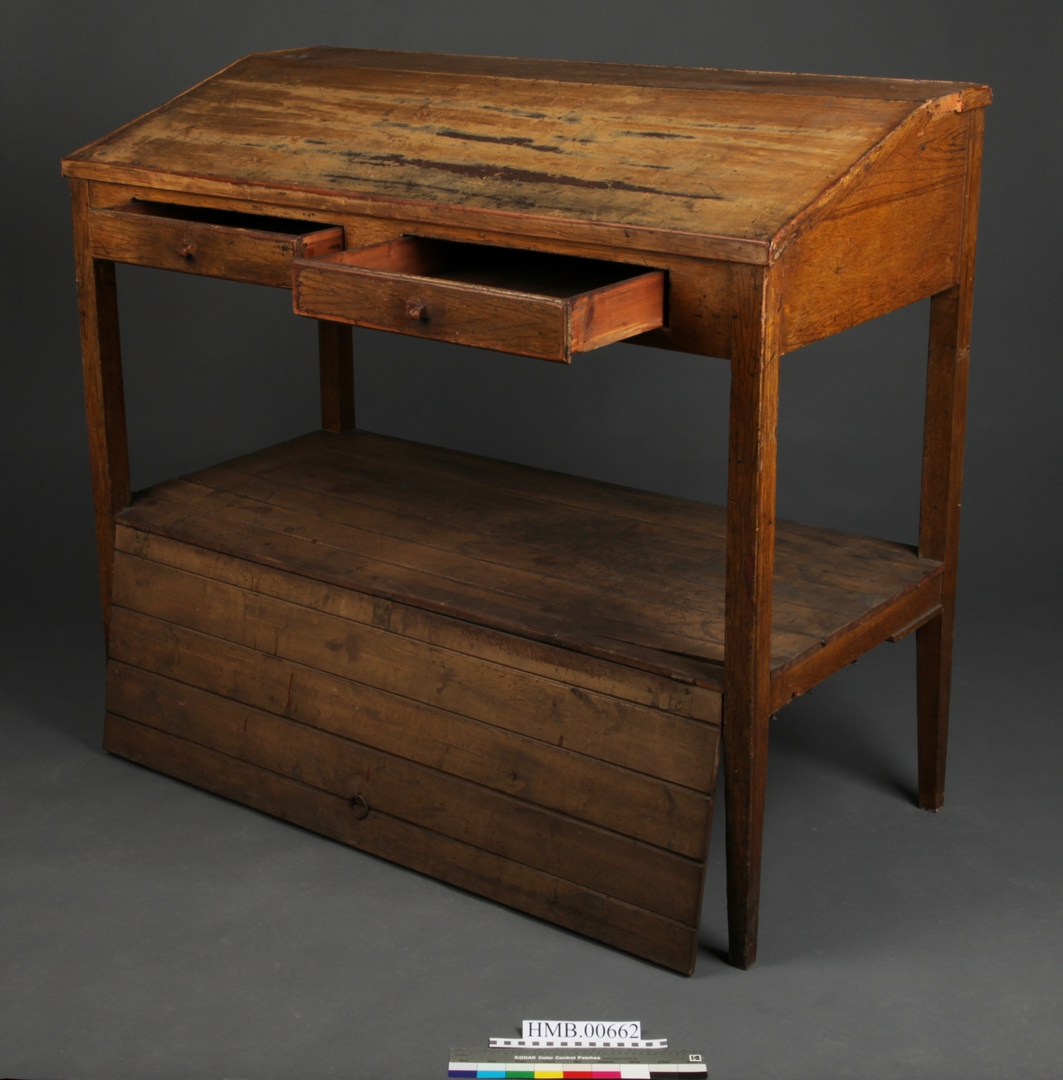 Rektangulært med skrånende skriveplate. To skuffer i front. Høye bein. Omtrent midt på beina er en hylleplate hvor det er hengslet en skapdør med en ring til håndtak. Skapdøren er hengslet i overkant og trekkes opp for å åpne.