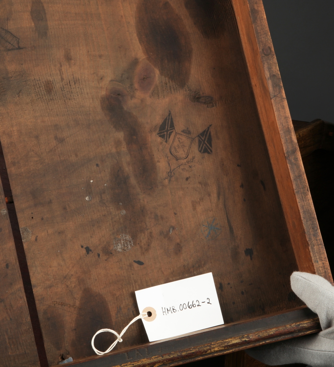 Rektangulært med skrånende skriveplate. To skuffer i front. Høye bein. Omtrent midt på beina er en hylleplate hvor det er hengslet en skapdør med en ring til håndtak. Skapdøren er hengslet i overkant og trekkes opp for å åpne.