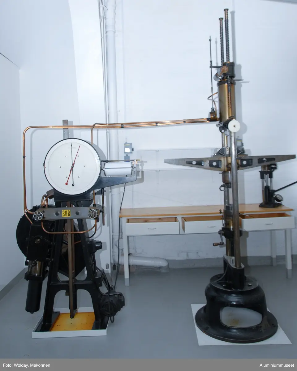 Strekkprøvemaskin for metaller.  Leverandør: Alfred J. Amsler & Co. Schweiz.  Datering: ca. 1925-1966. Teknikk: Hovedenhetene er støpt i stål og delvis maskinert.  Enkelte deler er fremstilt av valset stål og hydrauliske overføringer er kobber-rør.
Form: To stk. maskinenheter.  En betjeningsenhet og en strekkenhet som er forbundet med hydrauliske rør.
Tilstand: Maskinen er operativ, men har hydrauliske lekasjer.
Utfyllende opplysninger:
Betjeningsenheten består av en hydraulisk pumpe som via 2 stk. reguleringsventiler leverer olje til sylinderen for pendelmanometeret og til hovedsylinderen på toppen av strekkenheten.  Strekkprøvene plasseres i klemmebakkene på strekkenheten, blyanten settes på papirrullen og høyre kran på betjeningsenheten åpnes.  Etterhvert som trykket økes blir prøven strukket helt til brudd.  Under strekket slår pendelen ut tilsvarende trykket i hovedsylinderen og strekk-kraften kan til enhver tid avleses på skala-skiven.  Via et snorsystem fra overbjelken hvor øvre klemmbakke sitter, vil strekkforløpet bli avtegnet på en papir-rull på betjeningsenheten og strekkforløpet blir avtegnet slik at hele strekkforløpet og flytgrensen kan avleses.  Ved bruk avstenges hovedkranen umiddelbart og da vil den sorte pila umiddelbart gå tilbake til null-posisjon., men den røde pila blir stående igjen og viser bruddkraften.  Med den venstre kranen slippes hovedsylinderen ned til utgangsposisjon igjen.  Tykkelse og bredde på prøvestaven blir målt før prøvetaking og staven blir også risset med tynne paralelle strekker med 10 mm mellomrom.  Se risseapparat HAM.79.  Etter brudd blir prøvestaven satt sammen igjen og målt over en lengde på 5 riss med bruddet ca. midt i mellom. Den nye lengden i forhold til 50 mm viser forlengensen som blir utregnet i prosent.  Bruddfastheten er avlest bruddkraft delt på tverrsnitt.  Flytegrensen avleses av det tegende diagrammet på papir-rullen pluss 0,2%.  Maskinen kan maks. yte 10 tonn, men pga. unøyaktig avlesing ved tynne prøver blir pendelmanometeret innstilt ettter forventet bruddstyrke.  Maskinen har 4 innstillnger: Maks.: 1000 kg: Nest nederste hull. Uten lodd. Maks.: 2000 kg: Nest øverste hull. Uten lodd. Maks.: 5000 kg: Nederste hull.  Med Lodd. Maks.: 10000 kg.: Nest øverste hull. Med lodd.
Maskinen står i dag 08.01.2007 i psosisjon, 5000 kg.  Maskinen kan også brukes til trykkprøving, f.eks. sveiseprøver, ved å plassere prøven på rullebakkene oppe på strekk-bommen.  22.01.2008 Kjell Grette.