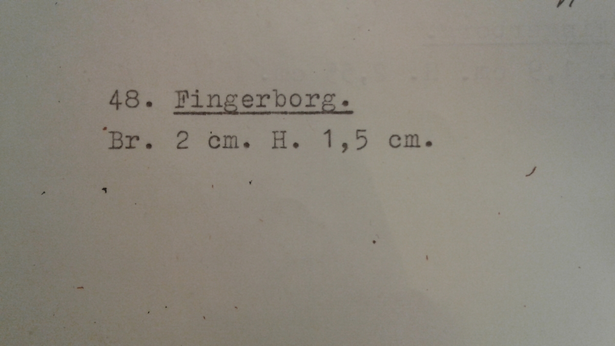 Svagt konisk fingerborg av brons med relativt glesa gropar. Nedtill är en slät rand med fabriksstämpel: H intryckt (se bild).