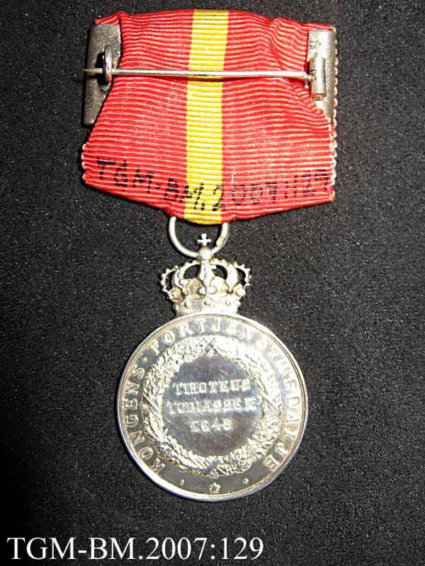 Medaljen ligger i en liten, flat, burgunderrød eske (original-etui) - preget med en forgylt krone på lokket. Silke og fløyel innvendig.