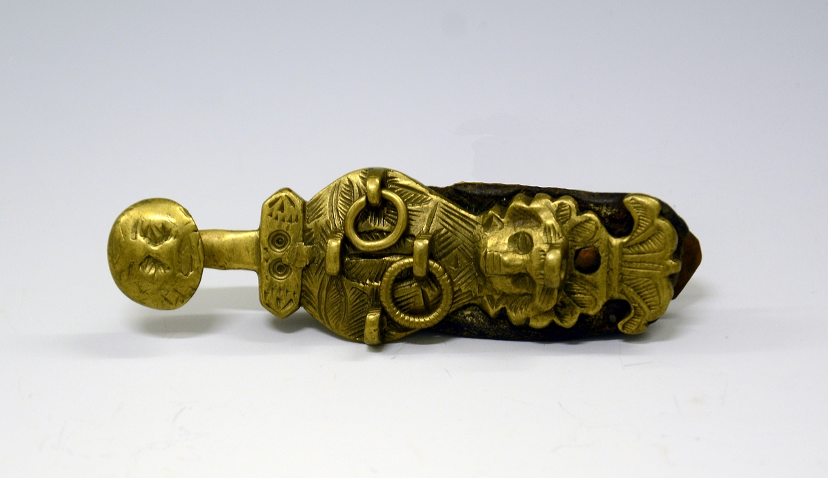 Slirehempe til belte og beltelås i messing. Fra protokollen: Bæltebeslag for tollekniver, av messing. Selve beslaget bestaar av a) 1 sprote (stolpelås eller beltelås) med paasatte 4-5 ringer og ornamenter i form av to masker.
b) En stolpe (slirehempe) med løkke paa. Fra stolpen gaar 2 par tvertagger, som gir plads til rembeslagene. Disse, der bestaar av 2 dobbeltbiøede plater ornerte med stjernepunkter, gaar rundt mellem tvertaggerne, og omslutter skindremmen som bokpermer. Beslaget har ringe og ornamenter.