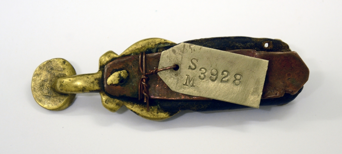 Slirehempe til belte og beltelås i messing. Fra protokollen: Bæltebeslag for tollekniver, av messing. Selve beslaget bestaar av a) 1 sprote (stolpelås eller beltelås) med paasatte 4-5 ringer og ornamenter i form av to masker.
b) En stolpe (slirehempe) med løkke paa. Fra stolpen gaar 2 par tvertagger, som gir plads til rembeslagene. Disse, der bestaar av 2 dobbeltbiøede plater ornerte med stjernepunkter, gaar rundt mellem tvertaggerne, og omslutter skindremmen som bokpermer. Beslaget har ringe og ornamenter.