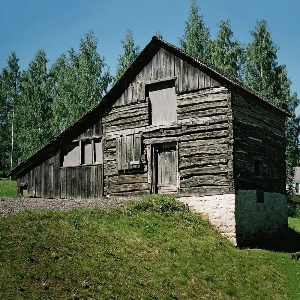 En enkel, laftet bygning i 1,5 etasjer med et tilbygg i reisverk på venstre side. Bygningen står på en gressdekket skråning. En hvitkalket grunnmur er synlig på høyre side. Bjørketrær i bakgrunnen.