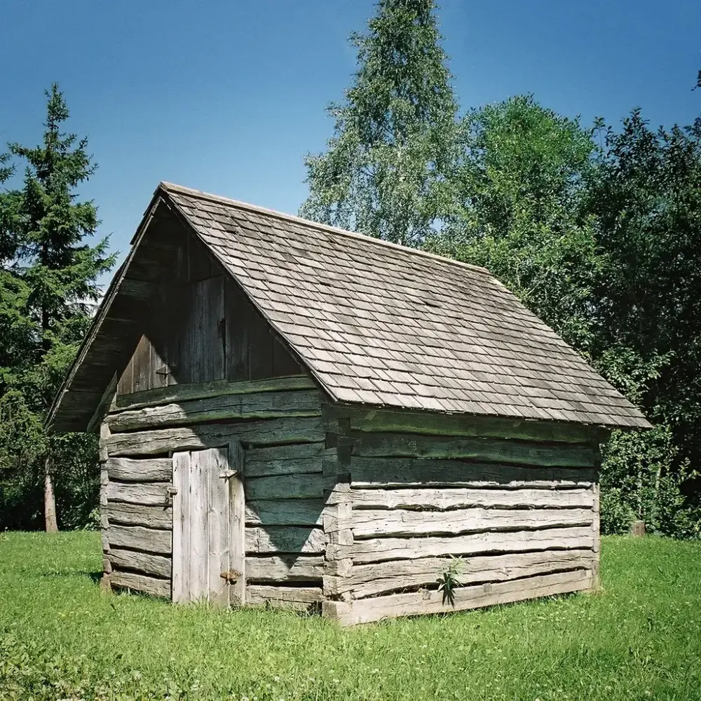 En liten, laftet bygning med en enkel plankedør i gavlveggen til venstre i bildet. Taket er tekket med trespon. Bygningen står grasbakke og har trær i bakgrunnen.