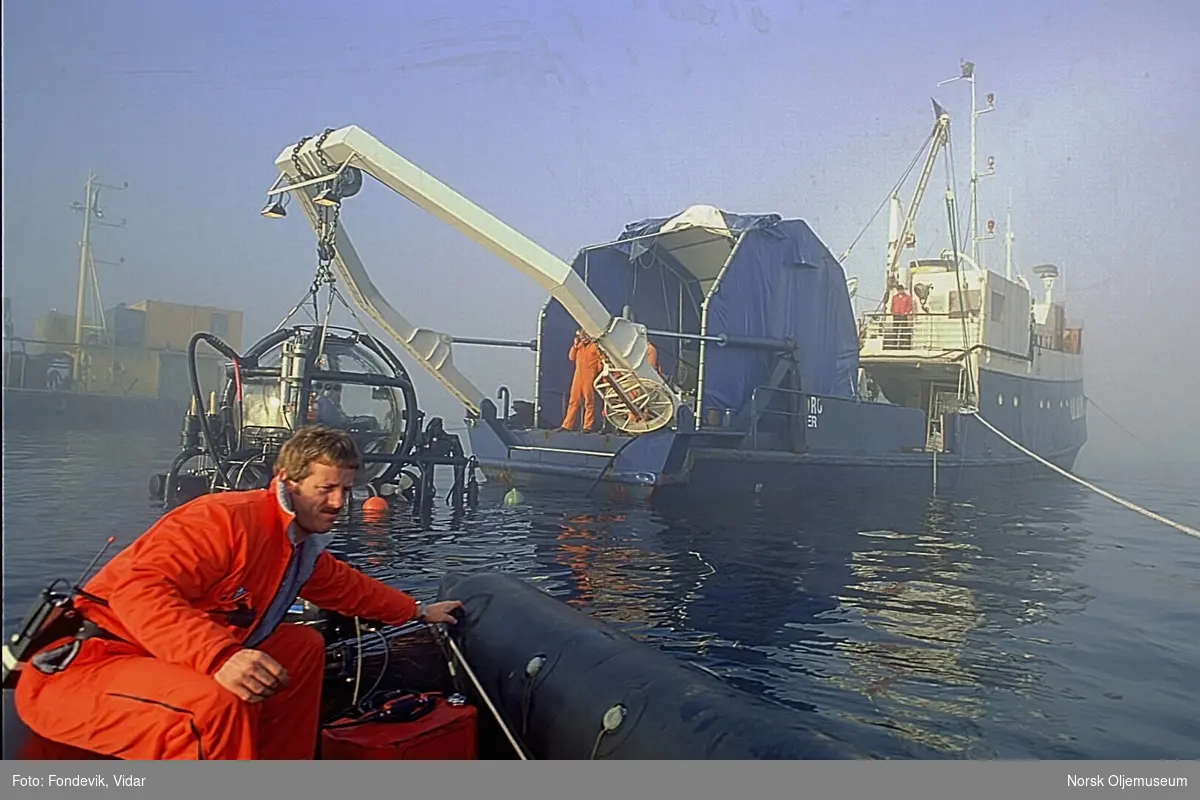 En mann kjører bort fra dykkerskipet "Strilborg" i en liten gummibåt.  Strilborg ble senere omdøpt til "Deepsea Inspector", og er moderskipet til miniubåten "Check Mate" som henger i kranbommen i akterenden av skipet.