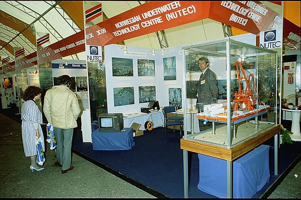 NUTEC sin stand på en messe. En person betjener standen.  En modell av "NUTEC Fjordbase" er plassert i en monter.