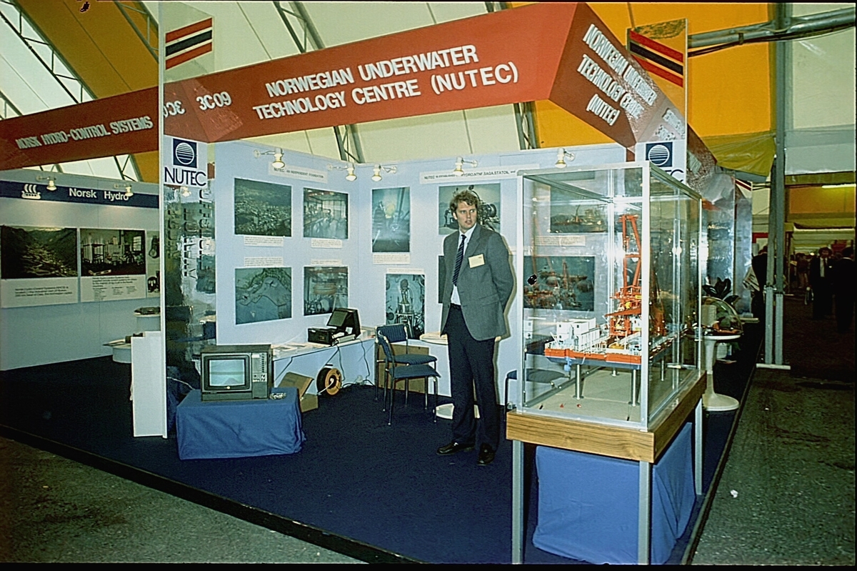 NUTEC sin stand på en messe. En person betjener standen.  En modell av "NUTEC Fjordbase" er plassert i en monter.