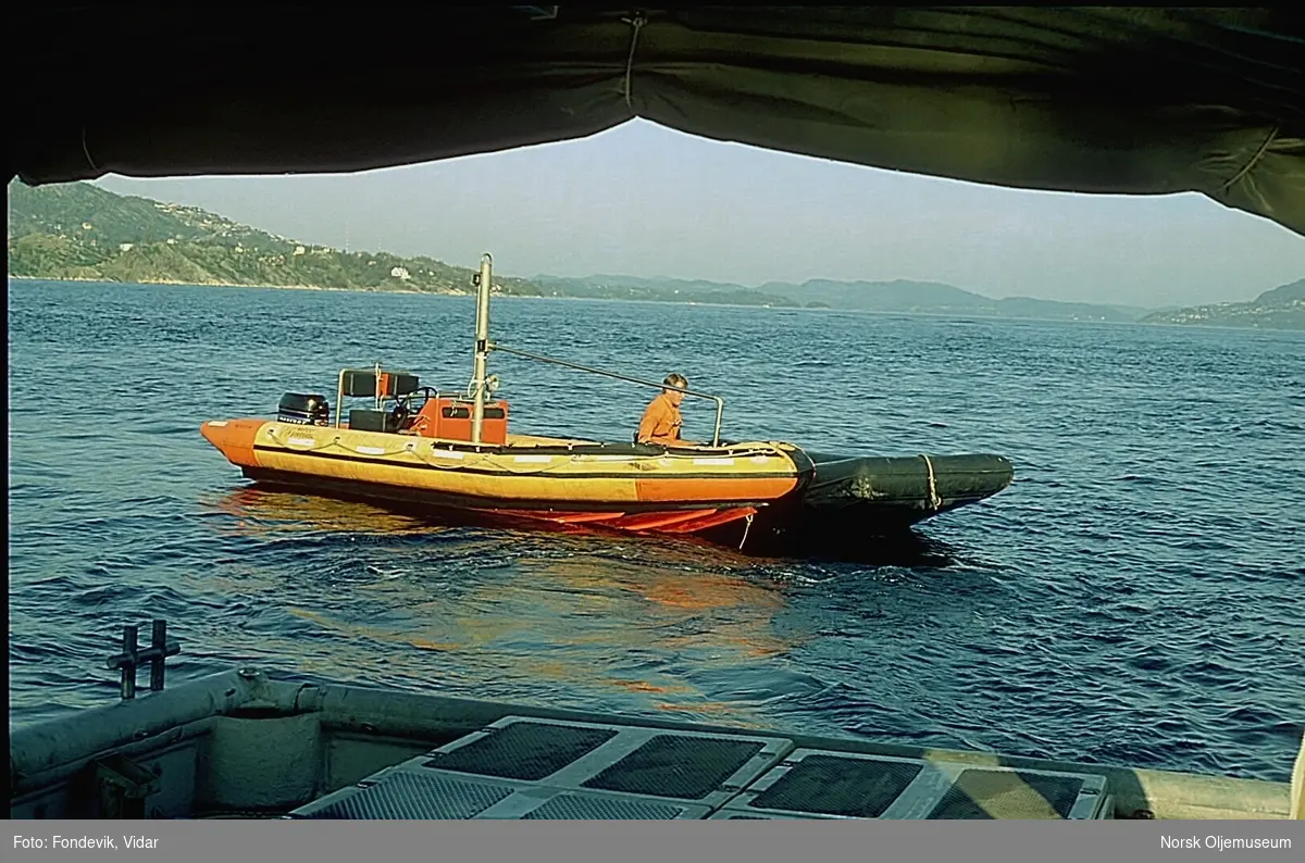 En mann sitter i en ribb på en fjord. Bildet er tatt fra akterenden på et skip.