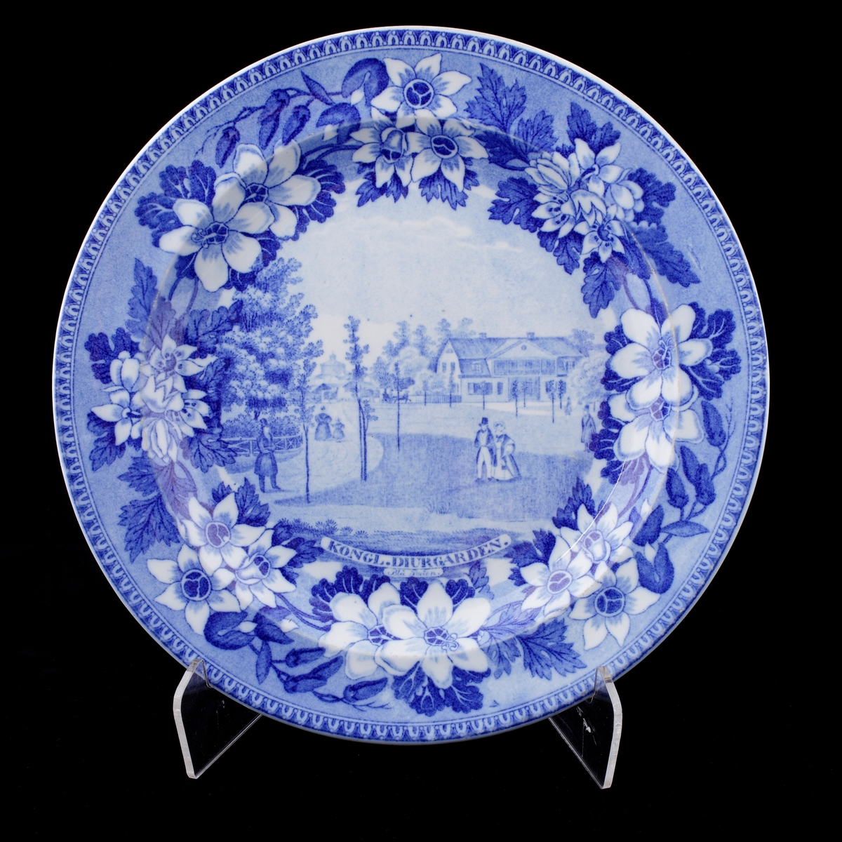 Tallrik av flintporslin.
Blåvit dekor (motiv från Kung. Djurgården), med blombård på brättet.