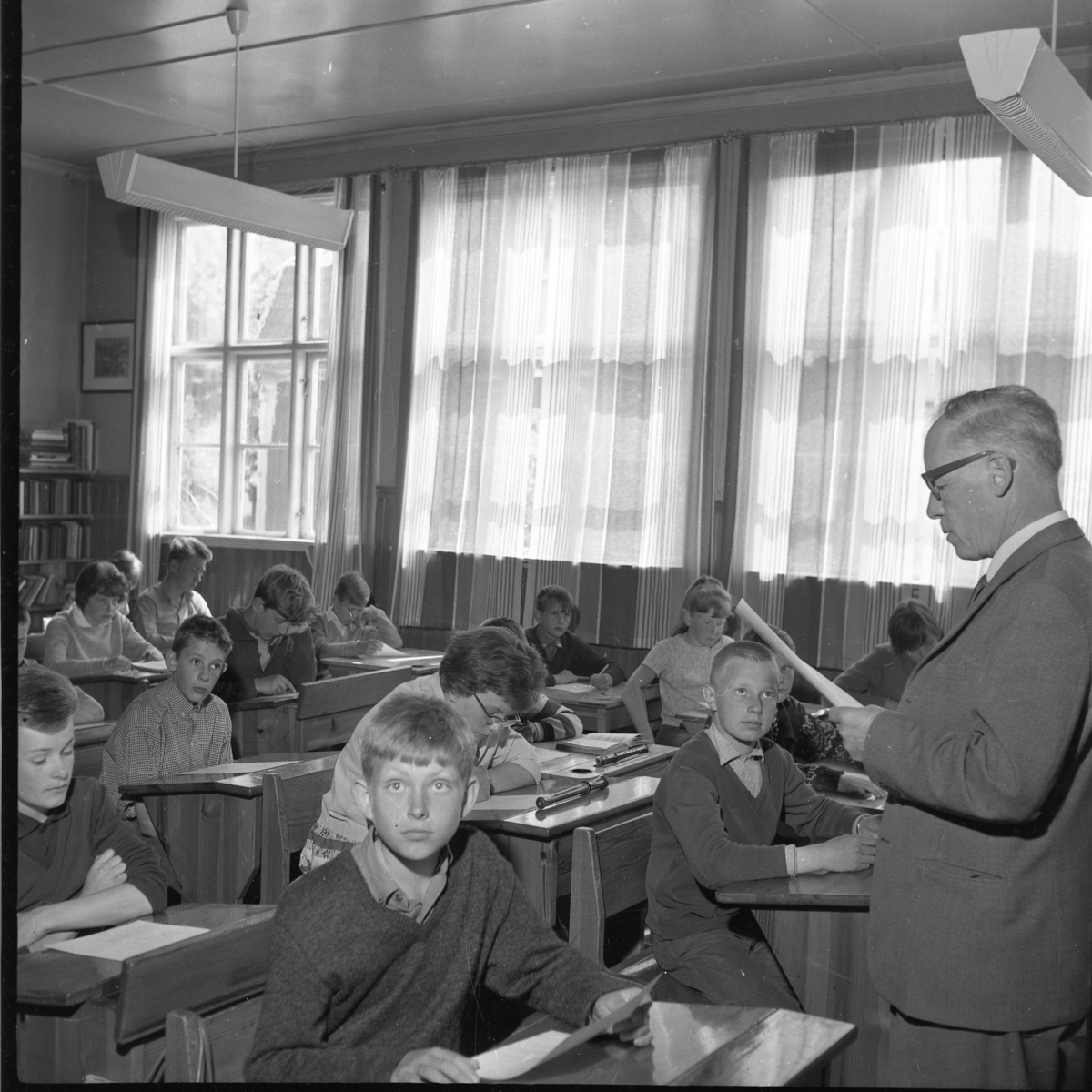 Sista lektionen i Uppgränna skola. Rektor Erland Skoog står framför eleverna, som sitter i sina bänkar, och läser i ett papper. Närmast i bild sitter Tommy Karlsson. Bakom honom Ronny Hermansson och intill honom Ros-Marie Jonsson med glasögon. Bakom henne sitter Peter Weidner i rutig skjorta.