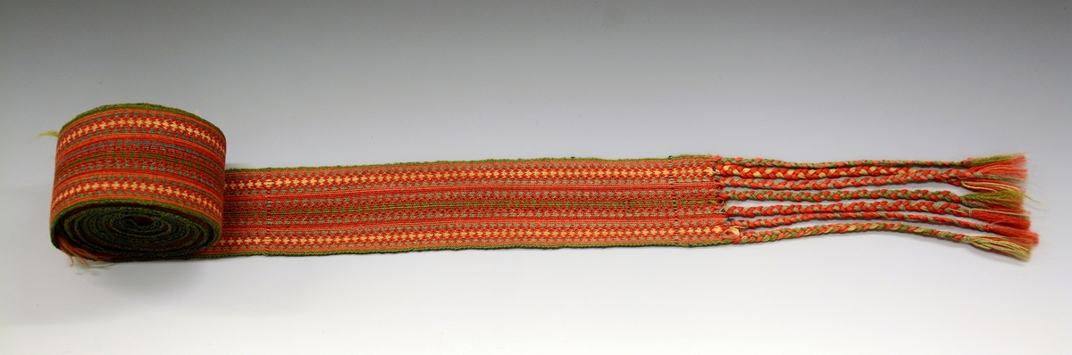 Belte til folkedrakt, brikkevevet. Frynser i den ene enden, de er flettet. Den andre enden er det renning uten avslutning.