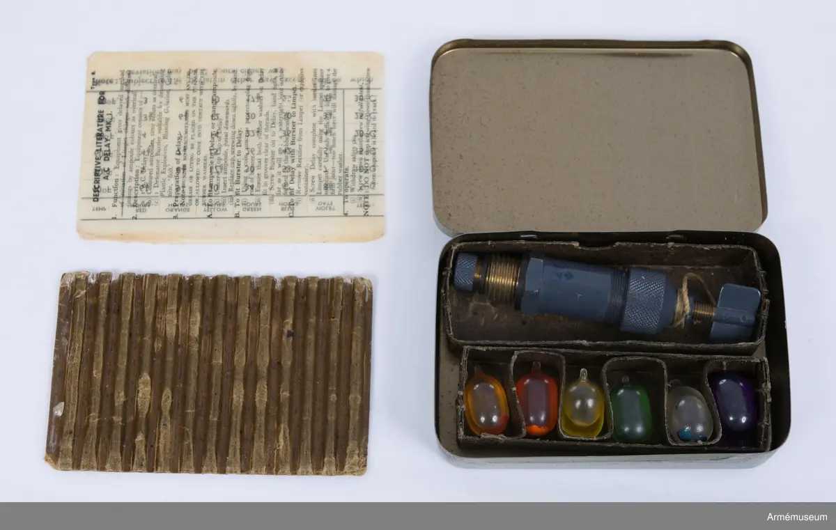 Grå plåtlåda med 6 ampuller innehållande acetonlösningar för olika tidsutlösningar.
Burster type 6.

A.C. Delay Mark I, TROPICAL PACK, Utlösare till fästmina:

With T/D Batch no (oläsligt), DATE 23 FEB 1945
BURSTER TYPE 6. TROPICAL PACK. A. 

Inklusive tändanordning och 6 st färgampuller med beskrivning. 
Acetone Celluloid
På instruktionen på insidan Type B
Vid 15 grader C fördröjs ampullernas utlösning med:
•	Röd 4 ½ time
•	Orange 7 ½ time
•	Gul 15 timmar
•	Grön 26 timmar
•	Blå 42 timmar
•	Violett 5 ½ dag
Notera att det kan ske en avvikelse på 15 % åt vardera hållet förutom Röd då avvikelsen kan vara 2 timmar åt båda hållen.