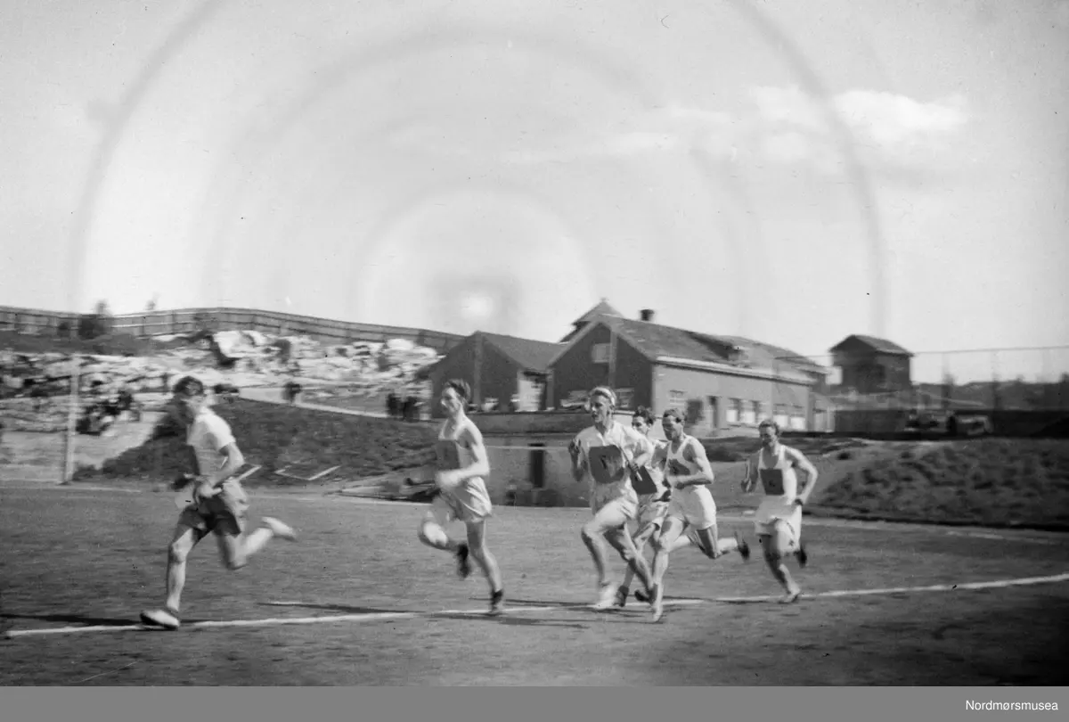 Dette er på Idrettsplassen i Kristiansund, Vi ser Mesterboligen der bak som står den dag idag (2018). Skimter også litt av museumsbyggene som ble flyttet ut til Knudtzondalen,  Dalen gård der de står i dag.  Til opplysning så er bildet speilvendt. (Info: Tor Aage Dyrseth Zdrinka.
2018)  løp, stafettløp. En fotosamling fra slekten Røv på Meisingset i Tingvoll kommune. Fotografiene kan trolig dateres mellom 1950-1960. Eier av originalmaterialet er Jan Harry Røv. Fra Nordmøre museums fotosamlinger.