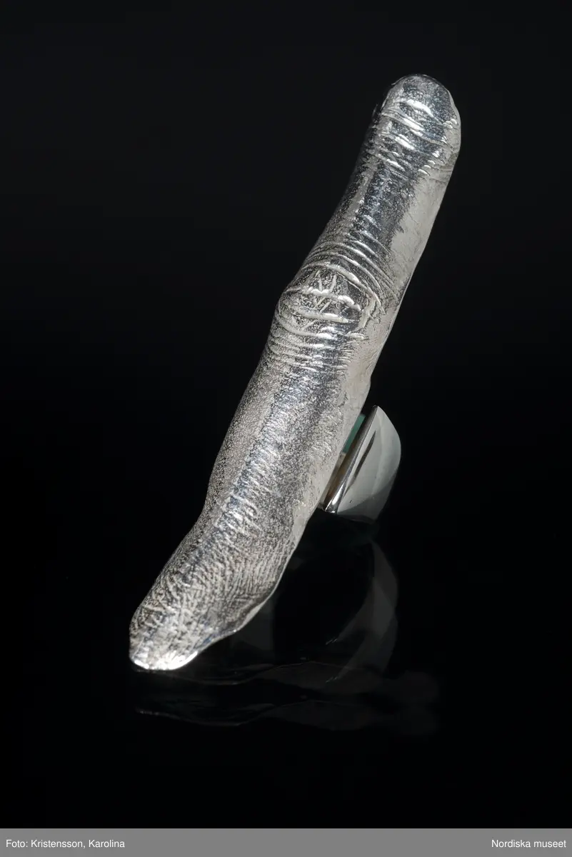 Ring av silver, överdel i form av avlång avgjutning av fingerrygg. Underdel bestående bred ring med slits på ena sidan. Undertill graverad "Cornelia".
/Leif Wallin 2018-02-09