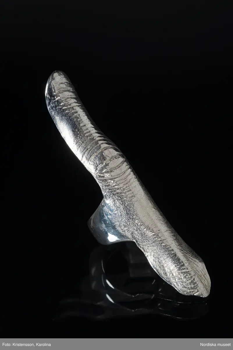 Ring av silver, överdel i form av avlång avgjutning av fingerrygg. Underdel bestående bred ring med slits på ena sidan. Undertill graverad "Cornelia".
/Leif Wallin 2018-02-09