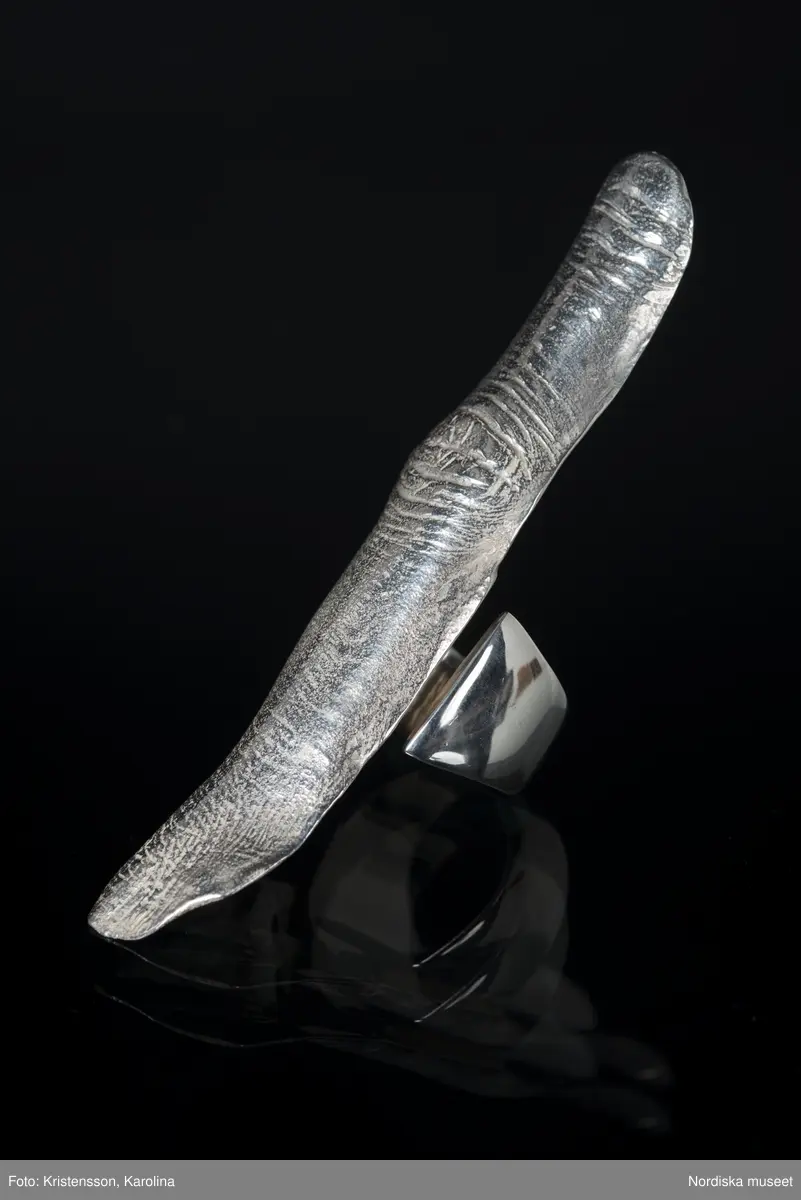 Ring av silver, överdel i form av avlång avgjutning av fingerrygg. Underdel bestående bred ring med slits på ena sidan. Undertill graverad "Cornelia".
/Leif Wallin 2018-02-09