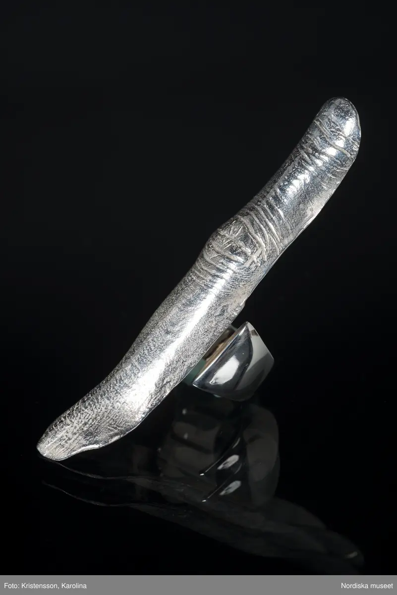 Ring av silver, överdel i form av avlång avgjutning av fingerrygg. Underdel bestående bred ring med slits på ena sidan. Undertill graverad "Cornelia".
/Leif Wallin 2018-02-09