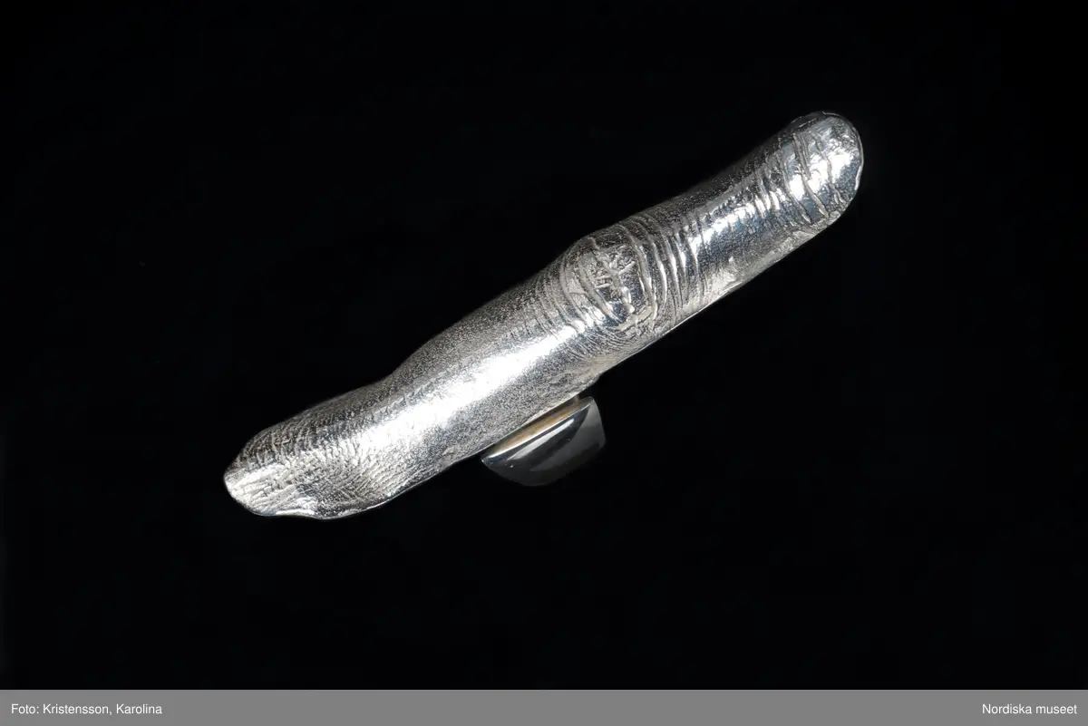 Ring av silver, överdel i form av avlång avgjutning av fingerrygg. Underdel bestående bred ring med slits på ena sidan. Undertill graverad "Cornelia".
/Leif Wallin 2018-02-09