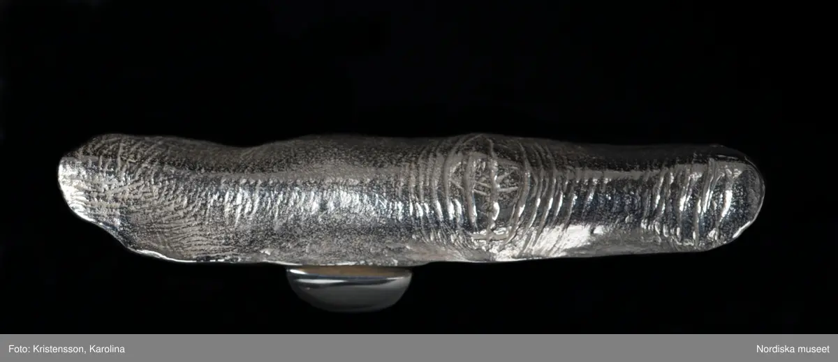 Ring av silver, överdel i form av avlång avgjutning av fingerrygg. Underdel bestående bred ring med slits på ena sidan. Undertill graverad "Cornelia".
/Leif Wallin 2018-02-09
