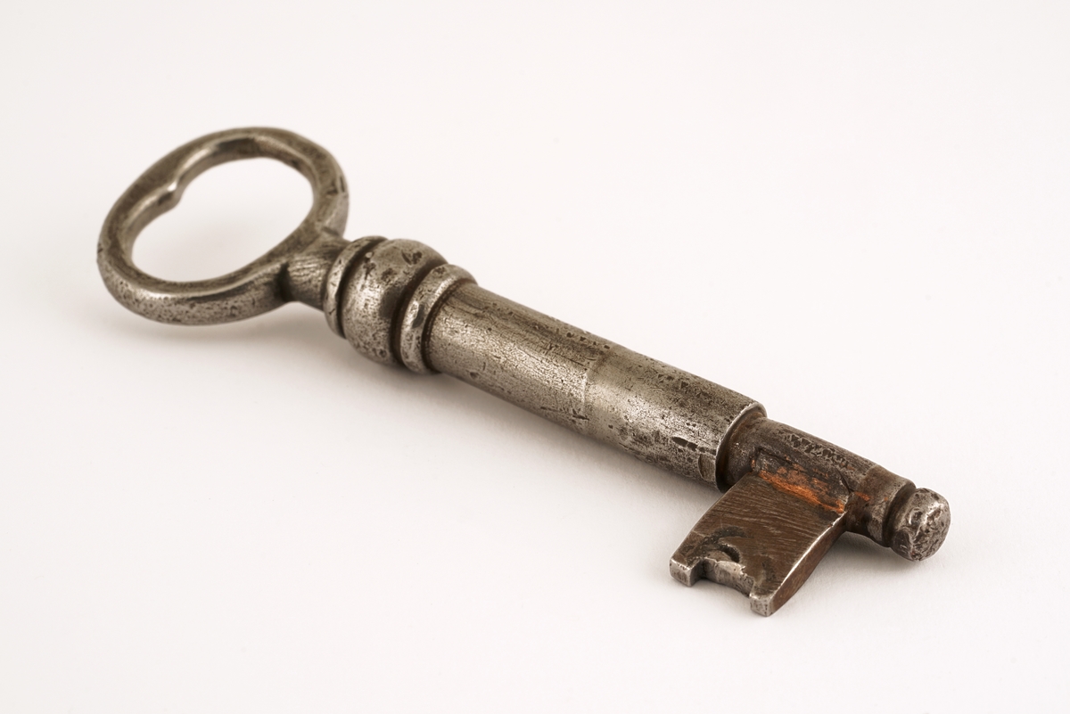 Dørlås for innmontering i dør av jern. Det medfølger tre monteringsbraketter, som selve låsen har vært montert på. Låskassen er rektangulær, med avrundede hjørner på den ene siden, og et større, flatt jern på den motsatte siden.Nøkkel følger med