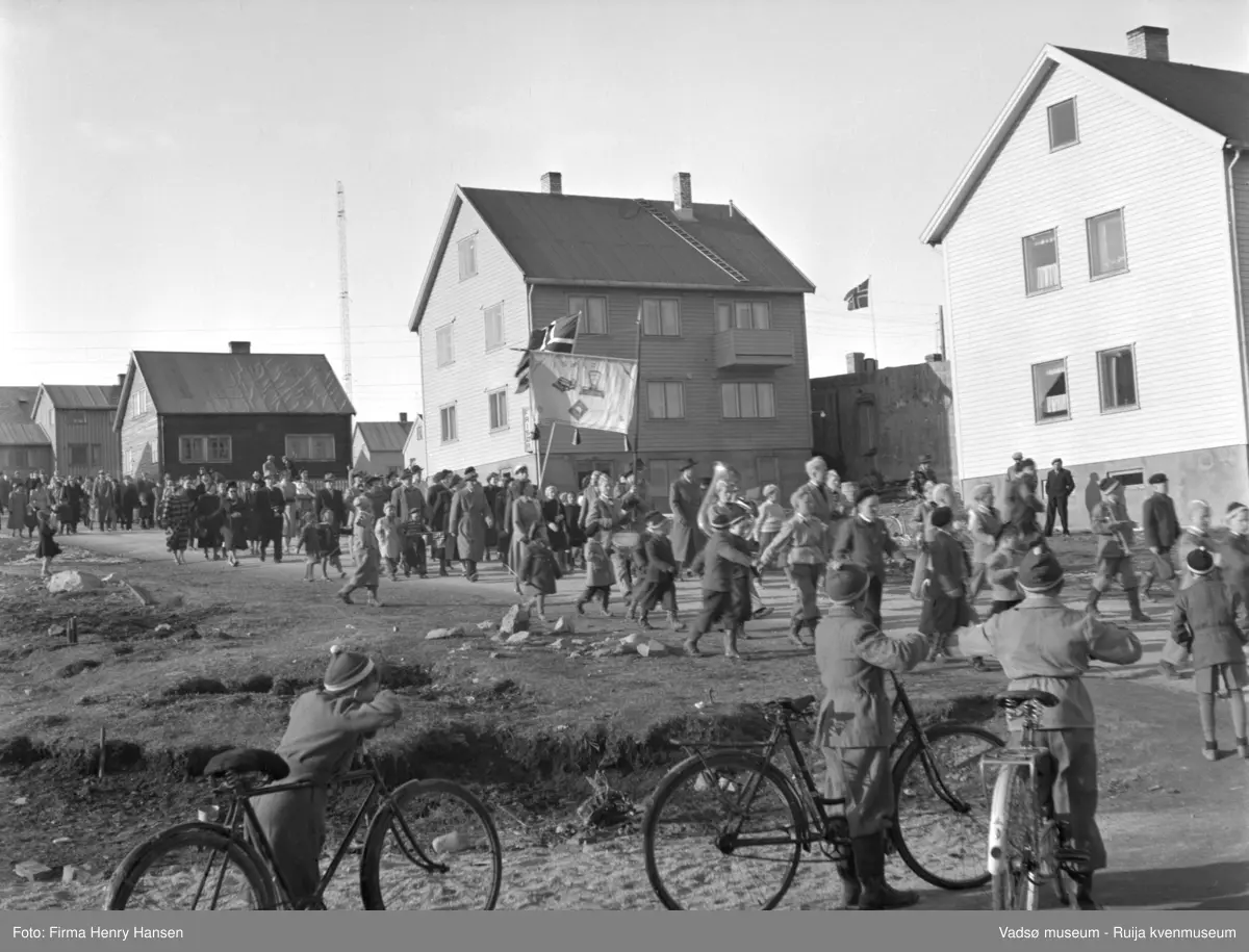 Vadsø 17 mai 1952. 17.mai-tog går mot øst, man ser en av kringkastingsmastene i bakgrunnen mot nordvest.. En fane har bilder og tekst, bl.a. en vekt og en skrivemaskin.