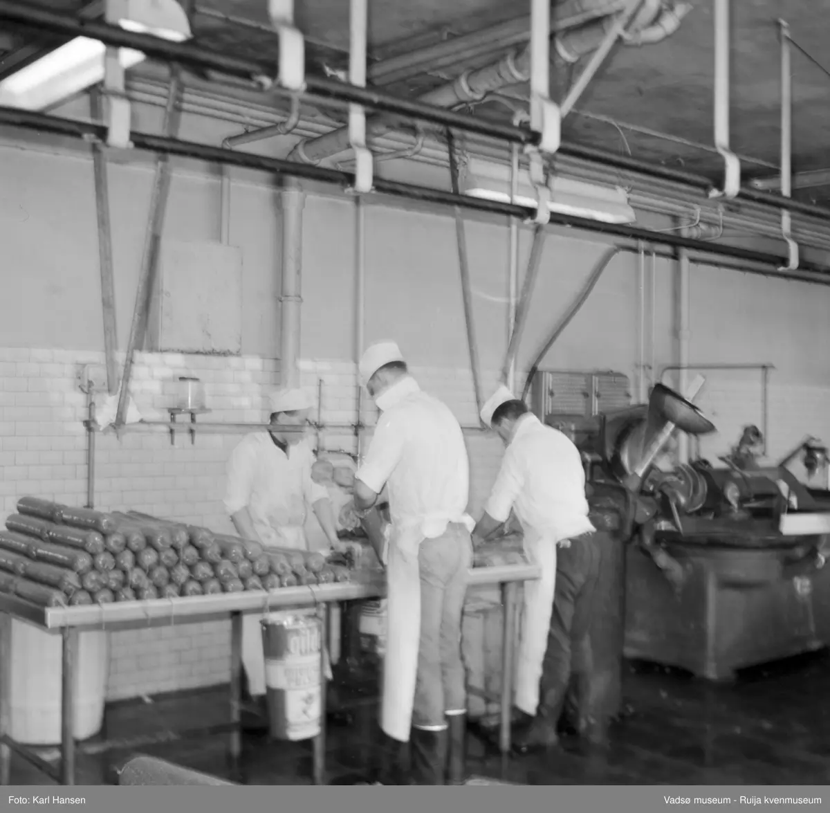 Nord-Norges salgslag Vadsø, 1964. Tre menn arbeider med spekepølseproduksjon i produksjonshallen. Fra venstre; Valter Kestilæ, Roy Aronsen og Ronny Johnsen.