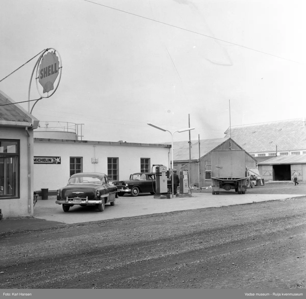 Vadsø sentrum 1959. Shell bensinstasjon og tankanlegg i Tollbugata. Reklameskilt, drivstoffpumper, to drosjer og en lastebil i forkant av bygningen. Til høyre i bildet ser vi skifertaket til Vadsø hermetikkfabrikk.