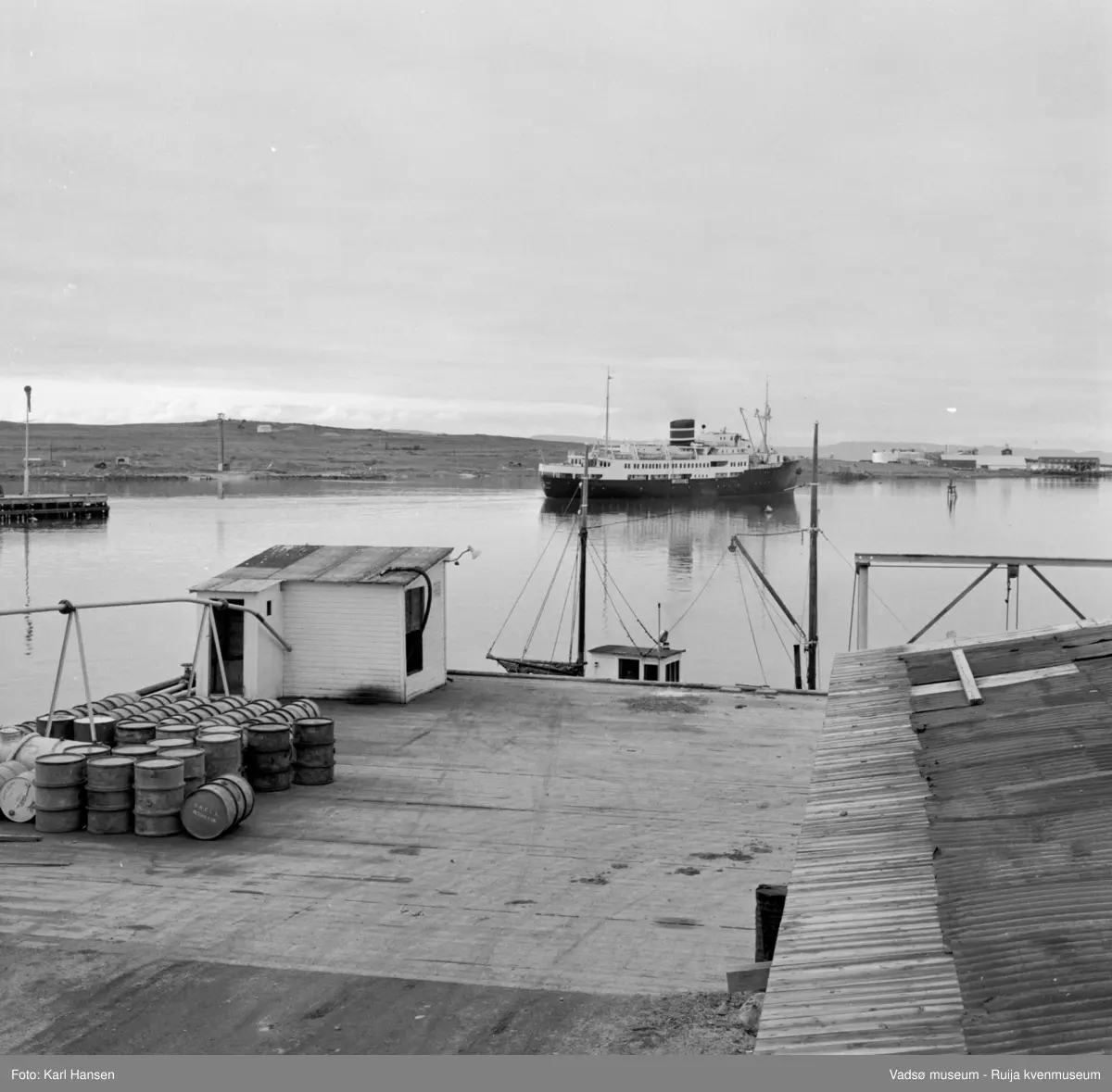 Vadsø sentrum 1959. Bildet tatt fra H.F.Esbensens "Shells" bensinstasjon og tankanlegg og anleggskai. Sjark ligger til kai. Hurtigruta på vei fra dampskipskaia. Vadsøya bak. Sildoljefabrikken ytterst på Øya.