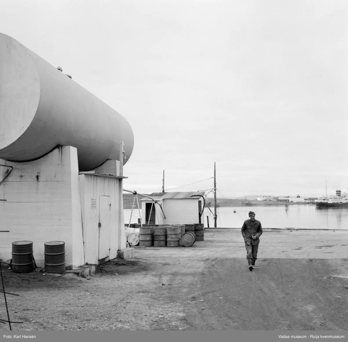 Vadsø sentrum 1959. Shells tankanlegg for drivstoff. Bildet er tatt ut mot havna med kaia mist i bildet. Tønner, fat med drivstoff lagret på kaia. På havna ser vi akterenden til hurtigruta og Vadsøya m/Vadsø Sildeoljefabrikk.