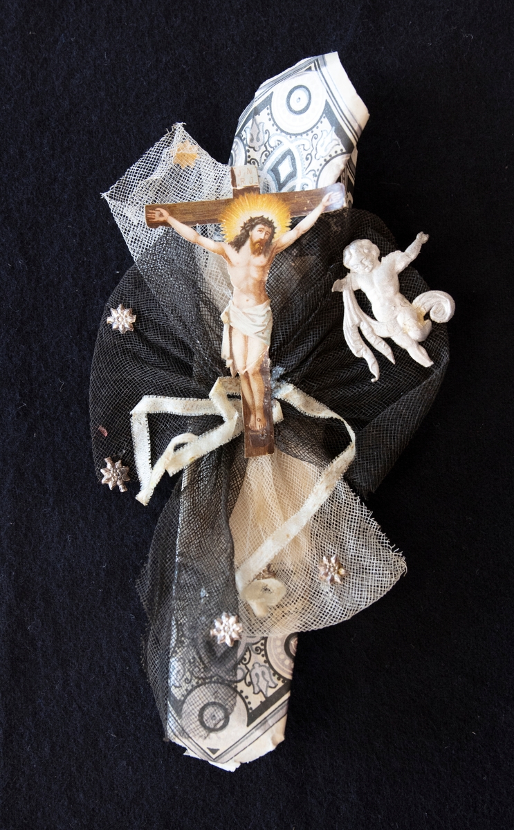 Begravningskaramell. I vitt mönstrat papper.  Två bokmärken på framsidan, en på Jesus på korset och en silverfärgad kerub. Rosetter på framsidan, en i svart och en i vitt. Rosetterna är försedd med silverstjärnor. Sötsaken är borttagen.

En av en samling karameller för dop-bröllop och begravningar.