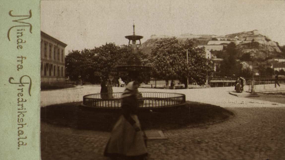 Nyttårskort. Fotografisk motiv. Kvinne foran fontene på et torg. Fredrikshald festning i bakgrunnen. Påtrykt tekst: "Minde fra Fredrikshald". "Olaf S. Madsen".
