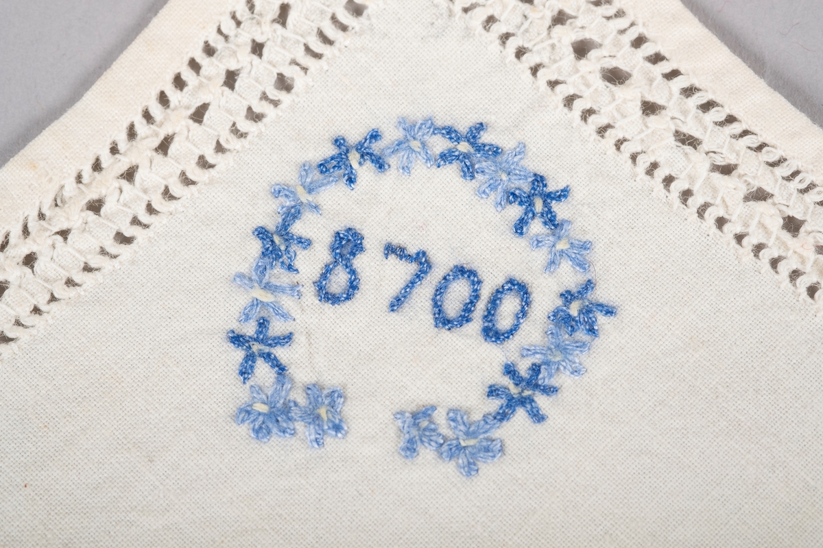 Hvit trekant lommetørkle med hullbroderier langs kanten og en brodert blå blomsterkrans rundt "8700", fangenummeret til Grete Wold.