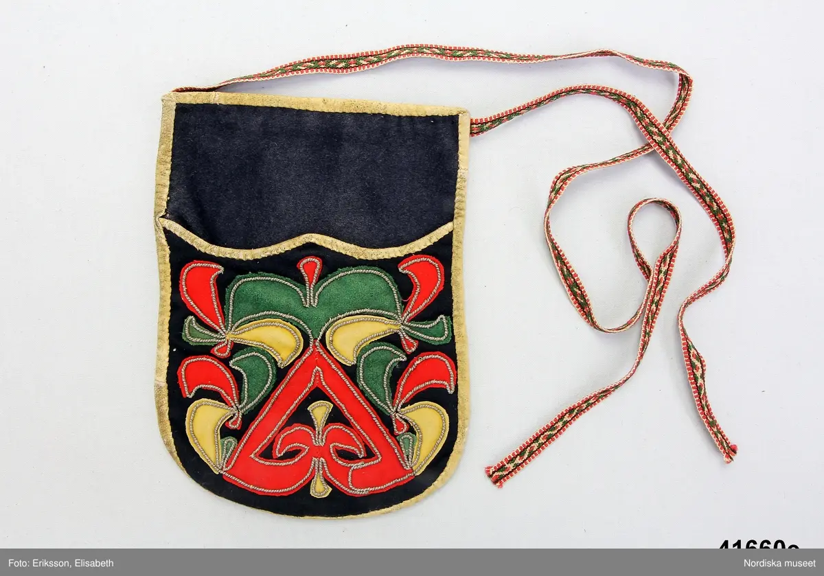 e. Kjolväska av mörkt blåsvart kläde.  kantad med ljust sämskskinn, på fickan applikationsbroderi med rött och gult kläde samt grön vadmal med tenntrådskonturer i läggsöm. Fickan insida av linnelärft, bakstycke av oblekt linnekypert.  Påsytt smalt midjeband av mönstrat halvylleband i vitt lingarn  med plockat mönster i grönt och rosa ullgarn. Avsett att knytas på vänster sida. 
I mycket gott skick
/Berit Eldvik 2012-02-14