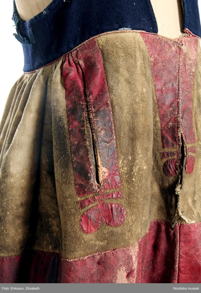 Huvudliggaren (år 1893):
"75386 Kvinnodräkt  fr. Herrestads hd. Skåne. a. Tröja af svart ylle med blåa sidenband. b. Skinnkjol med liv af blått ylle med blåa sidenband. c. Överdel med hålsöm. Märkt "K D L 1858". d. 'Smäck' af sidenband och guldband. e. List af brokigt bomullstyg med band och spetsar i guld och silver. Märkt '1840'. a. Banden sönder. Maläten b. Nött och något maläten."

c: Överdel. Material: Textil, halvlinne. Teknik: Tuskaft. Stopphålssöm, Plattsöm. Enkel utdragssöm. Korssöm. Knyppling. Längd: 35 cm. Längd: 66 cm - ärmlängd, totallängd, inkl spets. Vidd: 61 - ärmvidd. Vidd: 96 cm - midjevidd. Höjd: 8,5 cm - krage. Färg: Vit. Rosa - märkning. Brungul - hopdragssömmar och del av stopphålssöm på båltyget.
Överdel av vitt tuskaftat halvlinnetyg. Båltyget i ett stycke, foder över axelpartiet. Halsringning med 8,5 cm hög rak dubbelvikt krage. Kragen har en 2 cm bred bård i enkel utdagssöm i framkant. Broderat blommotiv  i plattsöm i övre kant fram. Sydda knapphål i sömmen mellan båltyg och krage. Sprund fram, 27,5 cm, invikt fåll, 0,8 cm. Smala brungula bårder i hopdragssöm i fållkant fram och tvärs över bålen där fodret slutar och bak.  1,5 cm bred bård i stopphålssöm över axelpartiet på båltyget. Broderad märkning ena sidan fram: "1858 KDV" [V- osäker], i ett motiv med monogram i rektangulära former, rött bomullsgarn, korssöm. Vidsydd ärm med ärmspjäll, stripade rynkor över axeln. Stripade rynkor mot 2,5 cm bred ärmlinning. Ärmlinningen består av en 2 cm bred bård i stopphålssöm, vit och brungul. 6,5 cm bred spets i framkant. Överdelen är handsydd.

Lästips: Svensson Sigfrid, (1935). Skånes Folkdräkter, en dräkthistorisk undersökning 1500 - 1900. Stockholm: Nordiska Museets Handlingar: 3.
/Inga-Lill Eliasson 2008-09-17

d. Bröstlapp, "smäck", höjd 20,5 cm, bredd 26 cm
fyrkantig på stomme av grå lumppapp med påsydda rader av sidenband mm. I mitten ett brett rödmönstrat sidenband omgivet av guldgaloner, längst ned en bit rödbottnad kattun och i övekanten ett blått och ett grönt enfärgat  sidenband, i nederkanten rester av ett äldre kattuntryck som verkar vara bortklippt.

Berit Eldvik mars 2005