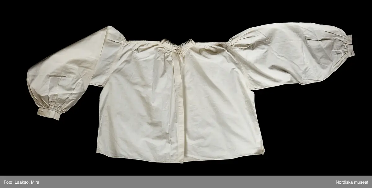 Huvudliggaren:
"Kvinnodräkt fr. Kåkinds hd, Västergötland.
b. Överdel av vit maskinvävd bomullslärft,  sydd som en blus med 2 framstycken och ett bakstycke, rynkade på axeln mot en smal axelslå, även rynkade mot 1,5 cm bred ståndkrage kantad med smal knypplad uddspets. Knäppt i halsen med liten vit pärlemorknapp och tränsat knapphål, övrig knäppning saknas. Vidsydd  mycket vid ärm med täta rynkor på kullen, rynkor nedtill mot 3 cm bred ärmlinning med sprund och 2 motstående knapphål. Maskinsydd.
Hör ihop med en hel rekonstruerad folkdräkt som bär spår av skärningen på 1890-talets modedräkt. Har tillhört frk. Lilli Zickerman, grundaren av Föreningen för Svensk Hemslöjd 1899. Hon kom från Skövde.
/Berit Eldvik 2010-09-21