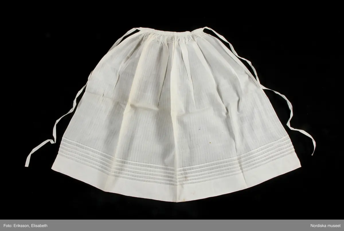 c: Förkläde. Material: Textil, bomull. Teknik: Manufakturvävning. Handsömnad. Längd: 77 cm. Bredd: 110 cm. Färg: Vit.
Förkläde i vitt fabriksvävt bomullstyg, sytt av 2 våder, smalrandigt, stärkt. Rynkningar mot midjelinning i tuskaftat bomullstyg, 1,5 cm bred, fastsydda bomullsband i sidorna. Uppvikt fåll nedtill, 6,5 cm, ovanför fåll, 5 lagda veck med ca 1 cm mellanrum, enkel utdragssöm i kanten av vecken.
/Inga-Lill Eliasson 2008-02-25

Lästips: Svensson Sigfrid, (1935). Skånes Folkdräkter, en dräkthistorisk undersökning 1500 - 1900. Stockholm: Nordiska Museets Handlingar: 3.
Jfr med förkläde 103 789d