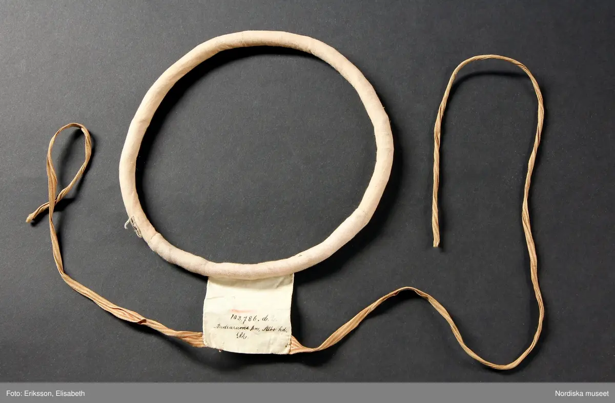 b. Brudtröja av svart vadmal,  Runt alla kanter bård av ytterst brunsvart sammetsband och innanför ett 4 cm brett svart sammetsband  med invävt bladmönster. På ärmarna ett svart slätt sammetsband med pålagd guldspets därefter siden band i rosa och grönt på botten av  svart  sammet och överst i bården en knypplad guldspets. 
c: Förkläde. Material: Textil, bomull. Teknik: Manufakturvävning. Handsömnad. Längd: 77 cm. Bredd: 110 cm. Färg: Vit.
Förkläde i vitt manufakturvävt bomullstyg, ihopsytt i mitten, smalrandigt, stärkt. Rynkningar mot midjelinning i tuskaftat bomullstyg, 1,5 cm bred, fastsydda bomullsband i sidorna. Uppvikt fåll nedtill, 6,5 cm, ovanför fåll, 5 lagda veck med ca 1 cm mellanrum, enkel utdragssöm i kanten av vecken.
/Inga-Lill Eliasson 2008-02-25

e: Krage. Längd: 47,5, cm - totallängd, bröststycke. Bredd: 33 cm - bröststycke. Höjd: 14 cm - kraghöjd, inkl spets. Material: Textil, bomull. Lingarn - spets. Bomullsgarn - broderi. Teknik: Tuskaft. Vitbroderi, Knyppling. Handsömnad.
Krage i tuskaftat vitt bomullstyg. Rektangulär duk, uppklippt halshål med fastsydd krage, 14 cm hög. Kragen har 3,5 cm bred fåll, enkel utdragssöm i kanten. Vitbroderi  i framkant,  plattsöm och kantsydda hål med infällt tyll, två sydda knapphål. Handknypplad spets fastsydd på långsida och korstsidor, 2,5 cm bred. Öppning fram, 24 cm långt sprund. Kragen är handsydd.
Lästips: Svensson Sigfrid, (1935). Skånes Folkdräkter, en dräkthistorisk undersökning 1500 - 1900. Stockholm: Nordiska Museets Handlingar: 3.
/Inga-Lill Eliasson 2008-02-19