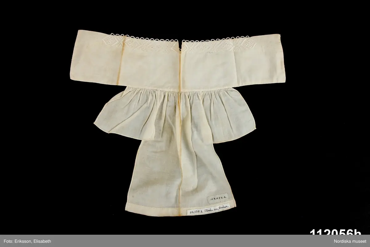 Mansdräkt från Floda sn bestående av:
a. blå vadmalsrock "blåtröja"
b. knäbyxor av sämskskinn, längd bak 90 cm, luckans br: 28 cm
c. väst av mörkblått kläde, längd bak 42 cm, fram 37 cm,
d. 1-2 ärmmuddar
e. strumpor
f. strumpeband, 
g. skjorta, 
h. nattkappa

Bilaga:
Finns ej.

Katalogkort:
g: "Skjorta. Längd: 102 cm. Axelbredd: 65 cm. Rakskuren. Hela framstycket av hellinne, ryggstycket av halvlinne. Ett 26. cm långt sprund framtill med ett veck, som döljer sprundet. 2 knapphål och 2 vita porslinsknappar. Skjortan rynkad vid kragen, vilken är 46 cm. lång och 9,5 cm. bred. Ett 9 cm långt sprund nedtill i varje sida. Ärmens längd 58 cm., vidd 49 cm. med spjäll. Ärmen nedtill rynkad vid den 3,7 cm. breda, med rätlinjig plattsöm broderade linningen. En vit porslinsknapp, ett knapphål."

b. KNÄBYXOR av sämskskinn, 2 fram-och 2 bakstycken midjegjord infodrad med svartrandig bomullskypert, sprund mitt bak, knäppt med 2 mässingsknappar, 4 hängselknappar, lucka av ovanligt slag med dubbla knapprader med 10 mässingsknappar, rikligt ornerade laskningar på luckan och på knäna med stjärnmönster, bensprund med 4 par hakar och hyskor av mässing, benlinningar för knäspännen, knälapp nedhängande.
c.VÄST 2 framstycken av mörkblått kläde, ett bakstycke av randigt bolstervarstyg i bounll i beige, rödbrunt, blått, gult, gråbrunt, gulbrunt och vitt. 2 iskurna fickor med 4,3 cm breda slåar. Dubbelknäppt med 16 svarta knappar och knapphål, en knapp har saknats är nu ersatt med annan knapp. Foder av kyprtat bomullstyg.
Berit Eldvik 2004

g: Längd: 102 cm. Längd: 58 cm - ärmlängd. Bredd: 46 cm - krage. Höjd: 9,5 cm - kraghöjd. Vidd: 112 cm. Vidd: 48 cm - ärmvidd.
Mansskjorta av kypertvävt linne i framstycket och kypertvävt halvlinne i bakstycket, övergången mellan hel- och halvlinne sker 6 cm nedanför axeln på framstycket. Traditionelll modell med dubbelvikt bålstycke, 9 cm långt sprund i sidorna. Rektangulärt halsspjäll med rynkningar mot krage. Dubbelvikt krage av fint helline, 9,5 cm hög, 2 porslinsknappar på insidan. Förslag med  sytt knapphål, längd: 26 cm, bredd: 10 cm. Vidsydd kypertvävd ärm i halvlinne med ärmspjäll. Rynkningar mot 3 cm bred ärmlinning med plattsömsbroderi i rombiska mindre former, vitt bomullsgarn, vit porslinsknapp och syttknapphål. Skjortan är handsydd.
/Inga-Lill Eliasson 2006-11-13

h: Nattkappa av tuskaftat vitt bomullstyg. Tre stycken, ett långt, 23 cm, och två kortare 9 cm på var sida, hopsydda och rynkade mot 9 cm hög krage som dubbelviks. Två cm bred broderad kantbård upptill, vinklar och små rombiska former i  rätlinjig plattsöm, vitt lingarn. Langetterade bågar utmed kanten. Nattkappan är handsydd, smala fållade kanter.
Anm: Gulnat.
/Inga-Lill Eliasson 2006-11-21