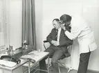 Interiör. Företagshälsovården (FHC). Läkarundersökning.
Personer: Patient: Adol Piatralla, efter namnbyte Adolf Hartenberger och Doktor Raxell.