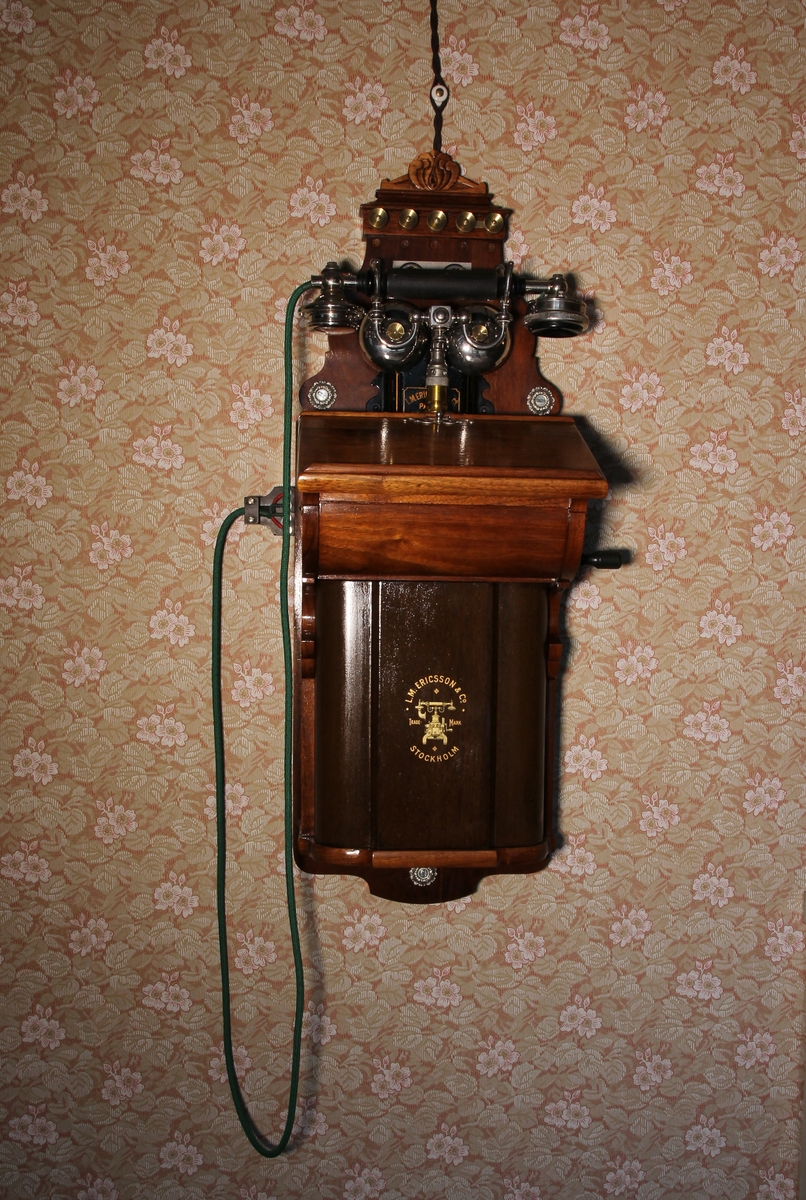 Telefon, väggapparat. Stående rektangulär med snedställd skrivplatta av brunbetsat trä. Klyka av vitmetall, lur och grönt telefonsnöre, Till höger vev. Kompletterad.
