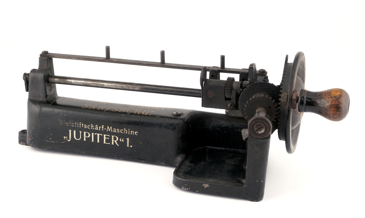 Blyantspissemaskin  av metall, sortlakkert med påskrift i gullfarge. Dreiet trehåndtak.
