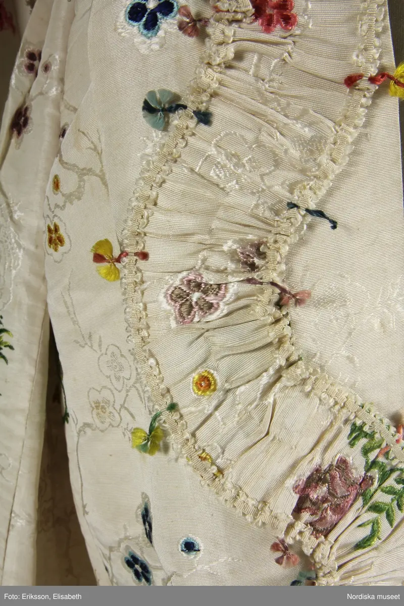 Brudklänning (robe), av broscherat, vitbottnat kinesiskt siden. Nygjord framvåd av fint vitt bomullstyg med kjolvolang nedtill av robens tyg. Två lösa garneringsbårder.