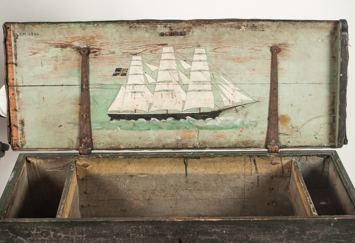 Sjömanskista, segelfartyg målat på insidan av locket.
Svartmålad och svart oljeduk ovanpå locket. Inuti finns en läddika t.h. samt ett öppet fack t.v. På lockets insida finns en fullriggare målad, den har seglen uppe och går från vänster till höger.