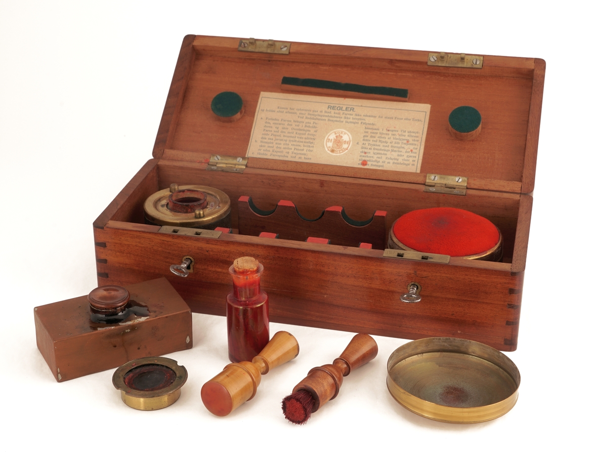 Trekasse med lokk med to låser med hver sin nøkkel. Inneholder to runde messingbeholdere, hvorav én med skrulokk, besatt med lær og med glassbeholder for fargestoff inni . Den andre med vanlig lokk, med stempelpute inni. Kassen inneholder i tillegg en børste med håndtak av tre,  ett stempelverktøy av tre, en glassflaske med kork inneholdene fargestoff, samt en rektangulær metallbeholder med skrukork.