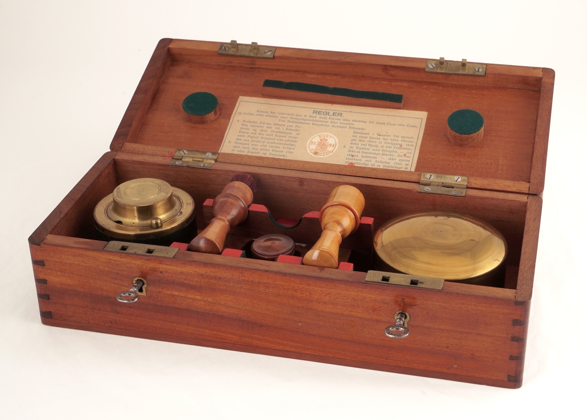 Trekasse med lokk med to låser med hver sin nøkkel. Inneholder to runde messingbeholdere, hvorav én med skrulokk, besatt med lær og med glassbeholder for fargestoff inni . Den andre med vanlig lokk, med stempelpute inni. Kassen inneholder i tillegg en børste med håndtak av tre,  ett stempelverktøy av tre, en glassflaske med kork inneholdene fargestoff, samt en rektangulær metallbeholder med skrukork.